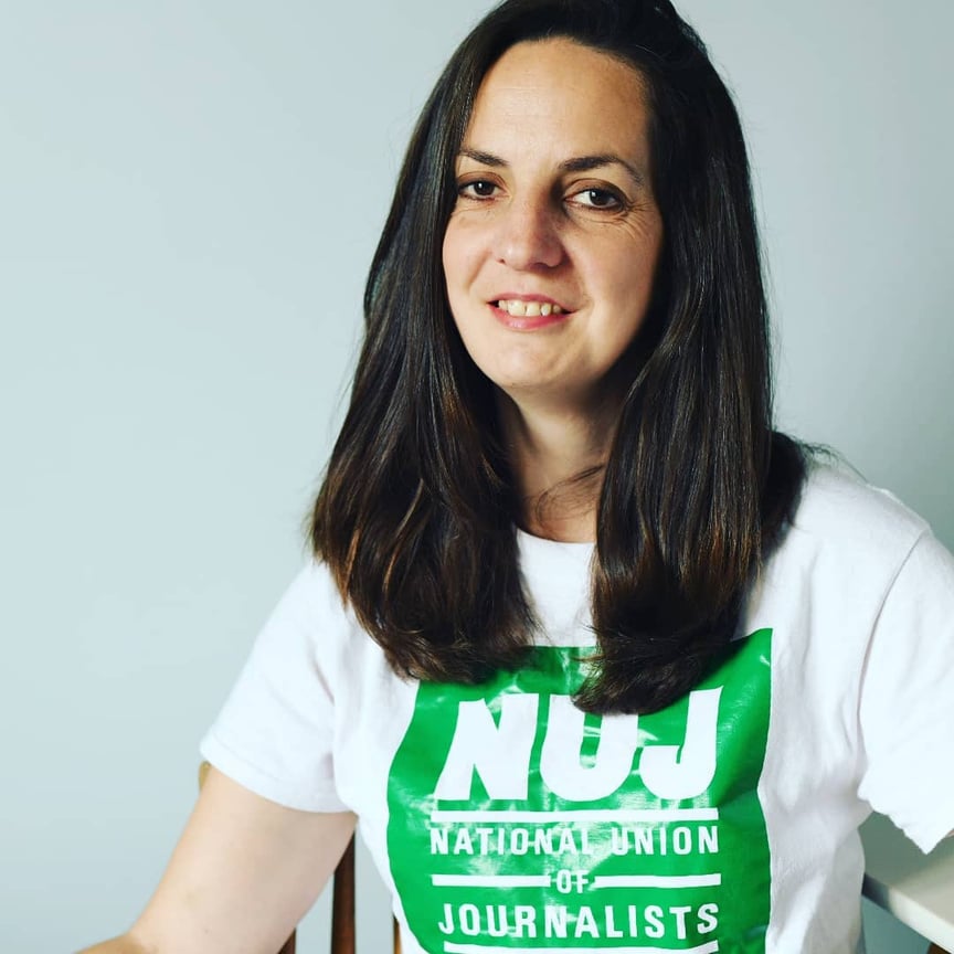 natasha, with dark brown hair past her shoulders wearing a white t shirt with a large green NUJ logo on the front. 