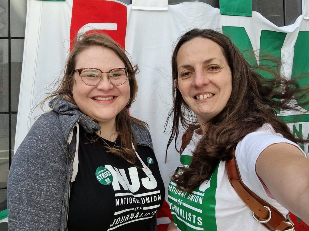 young member Polly on the left with natasha on the right, both wearing NUJ t-shirts and strike stickers with an NUJ banner in the background on the picket line in Cardiff