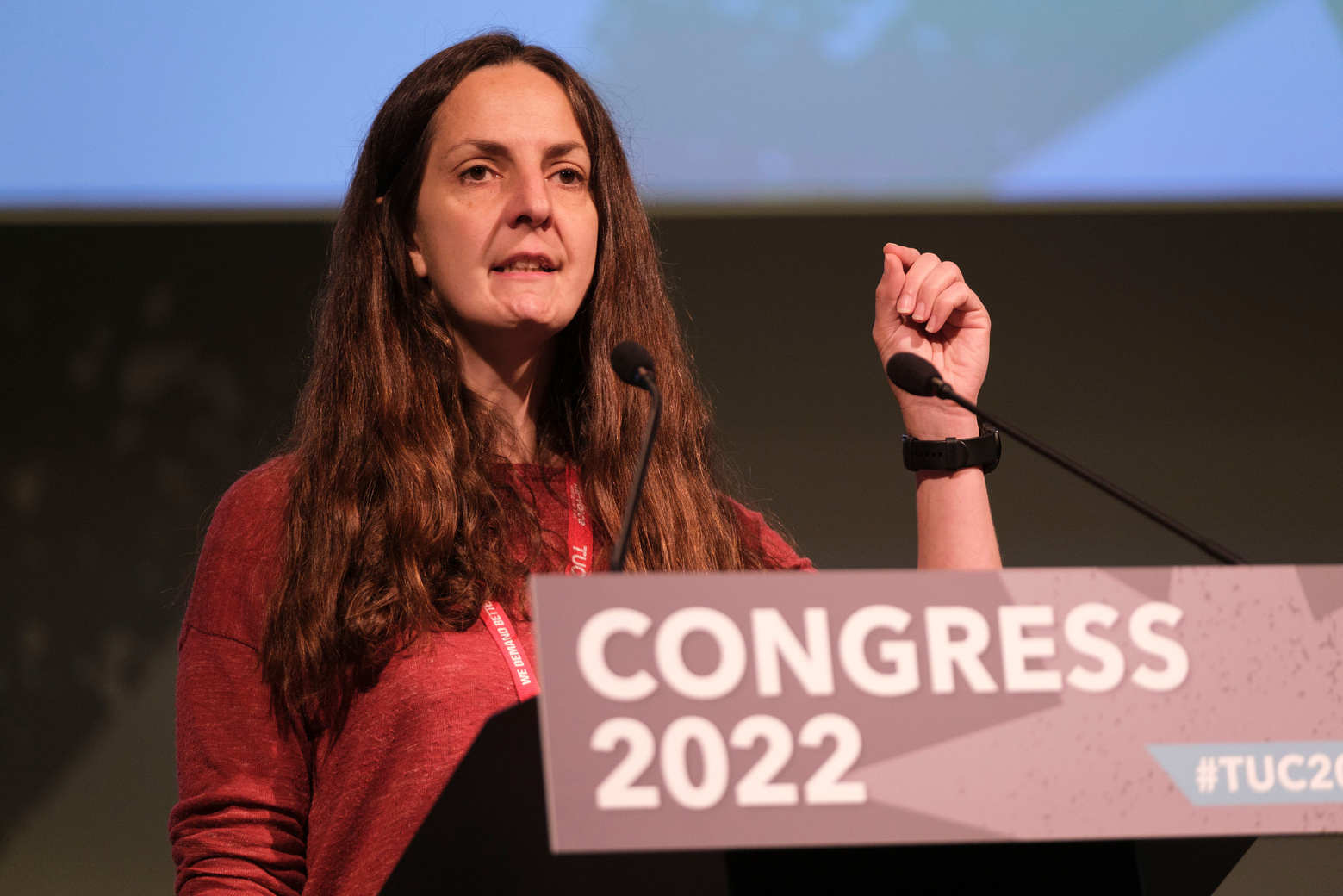 natasha with long brown hair and a red top with her fist in the air giving a speech at the podium at congress 2022