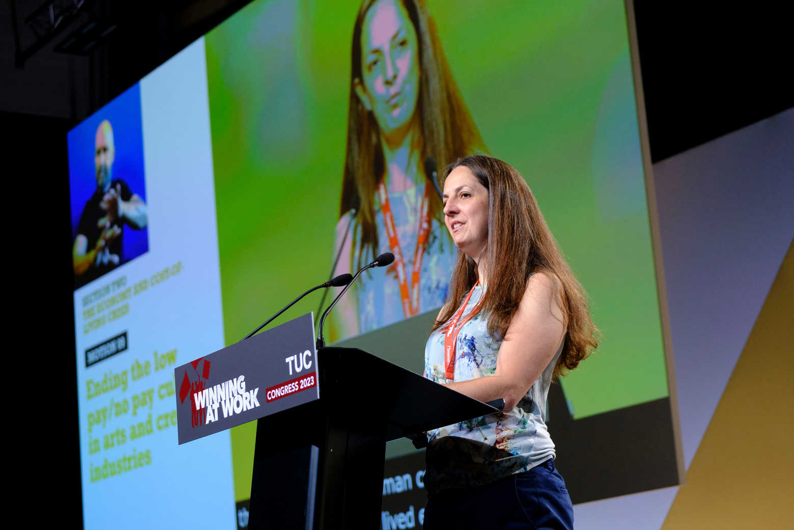 a side angle of natasha speaking from the podium, on a large stage. On the front of the podium, reads the slogan winning at work. in the background is a large screen showing natasha and the motion title, ending the low pay no pay culture in the arts